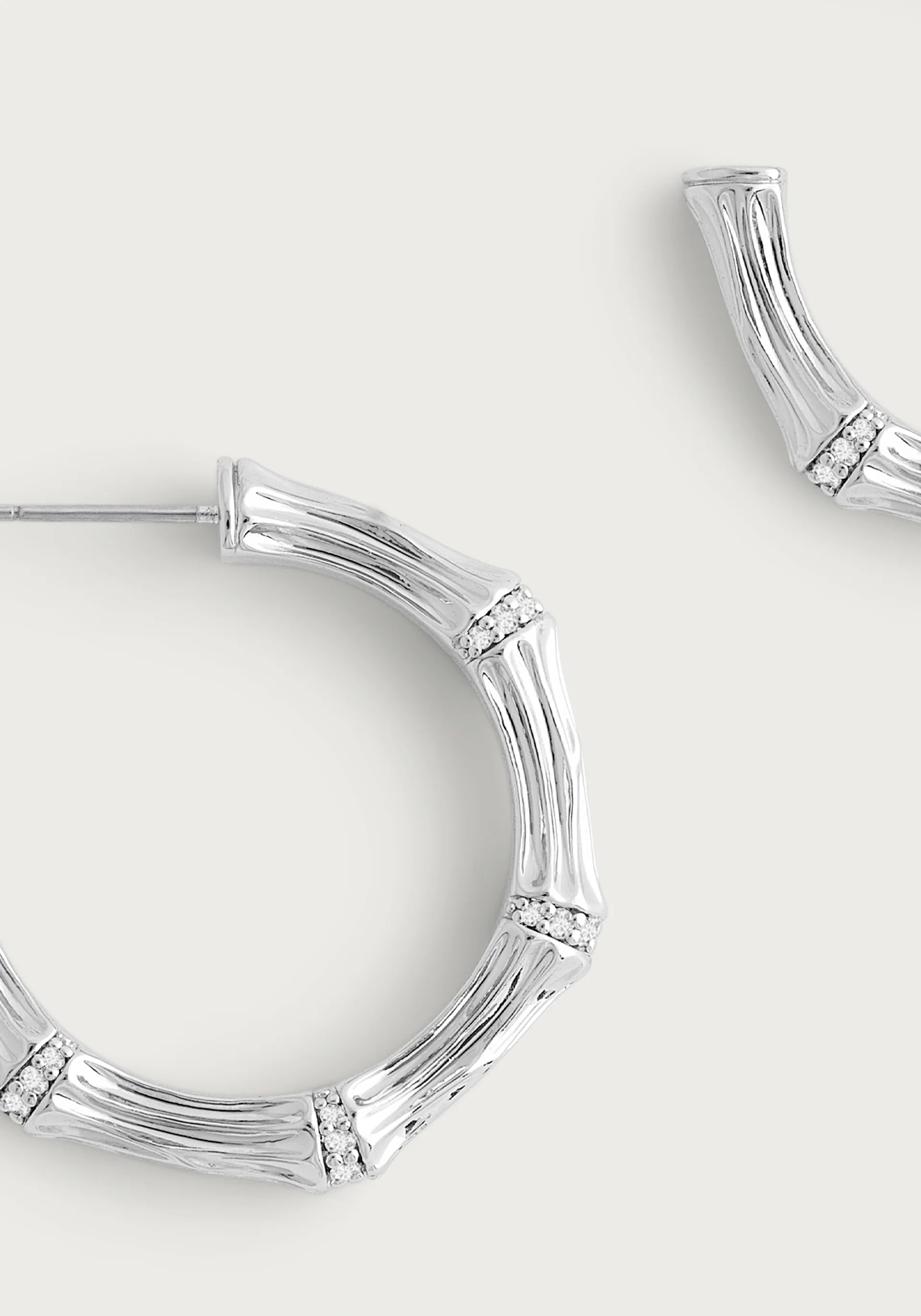 A close-up of the Bamboo Medium Hoop Earrings - Silver by Anabel Aram. Made from sterling silver plated brass, these earrings mimic bamboo segments with a textured finish and small sparkling stones at each joint, partially visible against a light background.