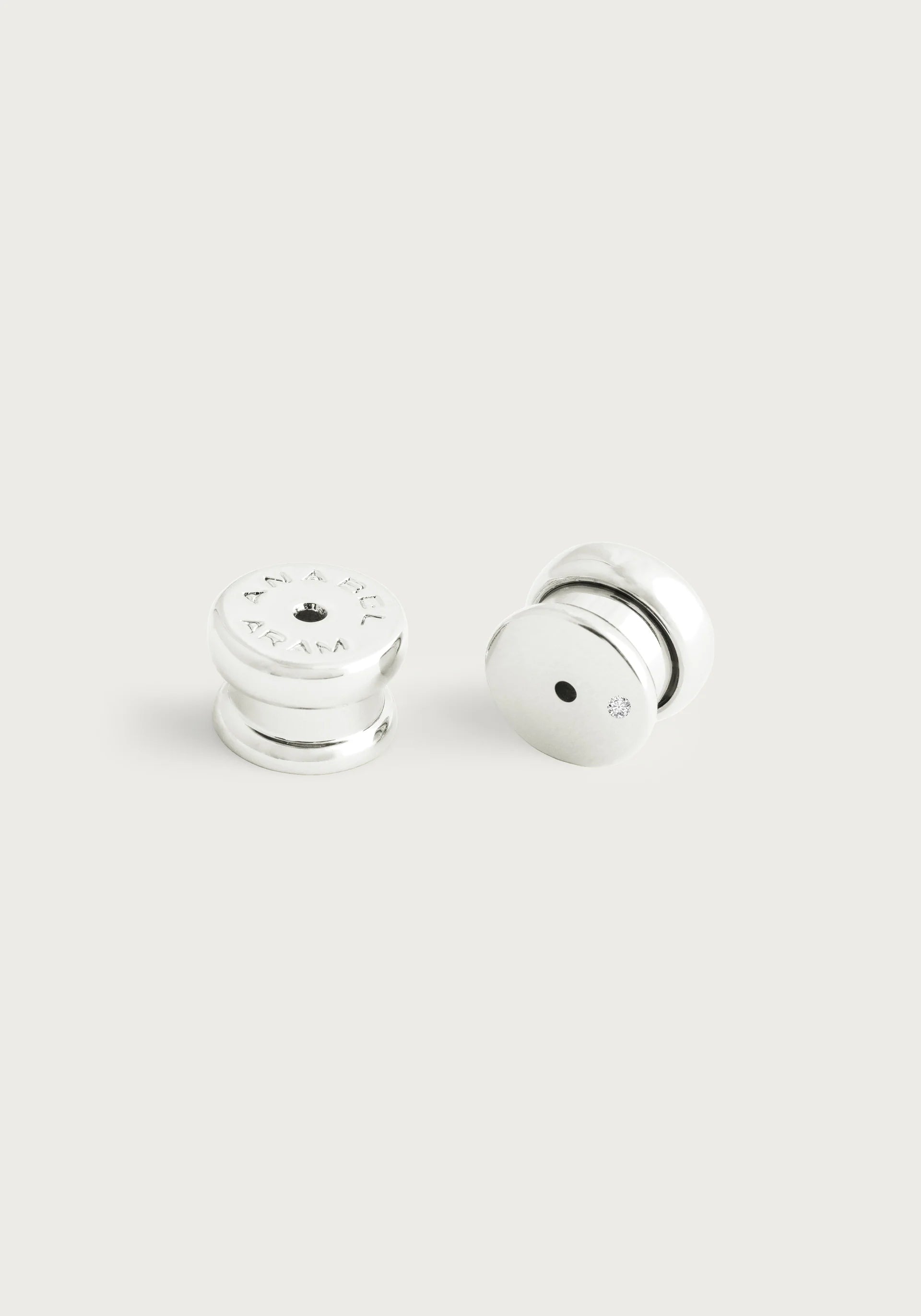 A pair of Bamboo Medium Hoop Earrings - Silver set on a light gray background. Crafted from sterling silver plated brass, one features an engraved logo, the other a small black dot. Their smooth, shiny finish exudes simple and modern elegance.