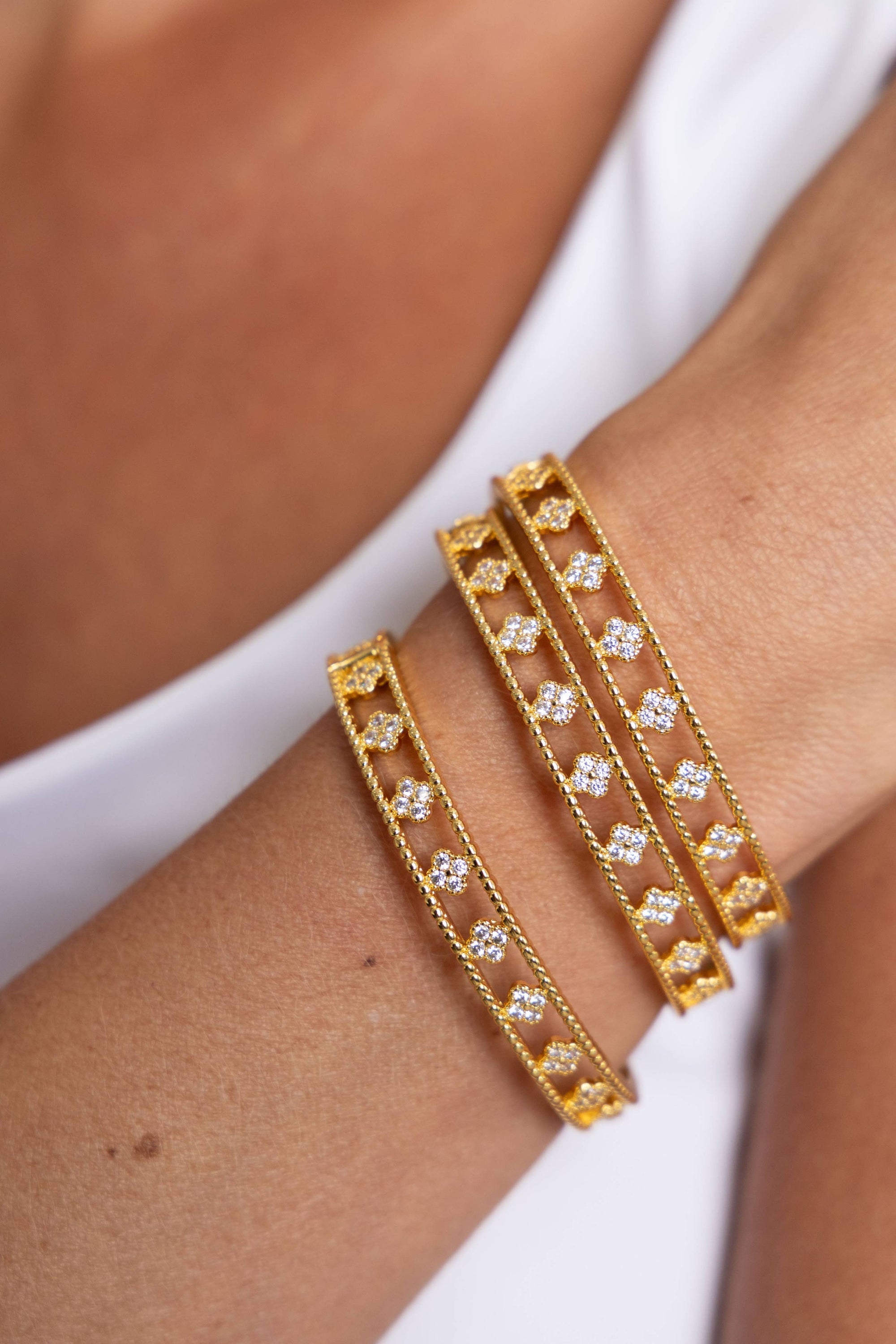 A close-up of a person's forearm displays three hypoallergenic Gracie CZ Clover Bangles, each crafted with 18k gold plating and featuring small cut-out heart shapes. Embellished with shimmering clear gemstones, these bracelets beautifully contrast against the simple backdrop of a white sleeveless top.