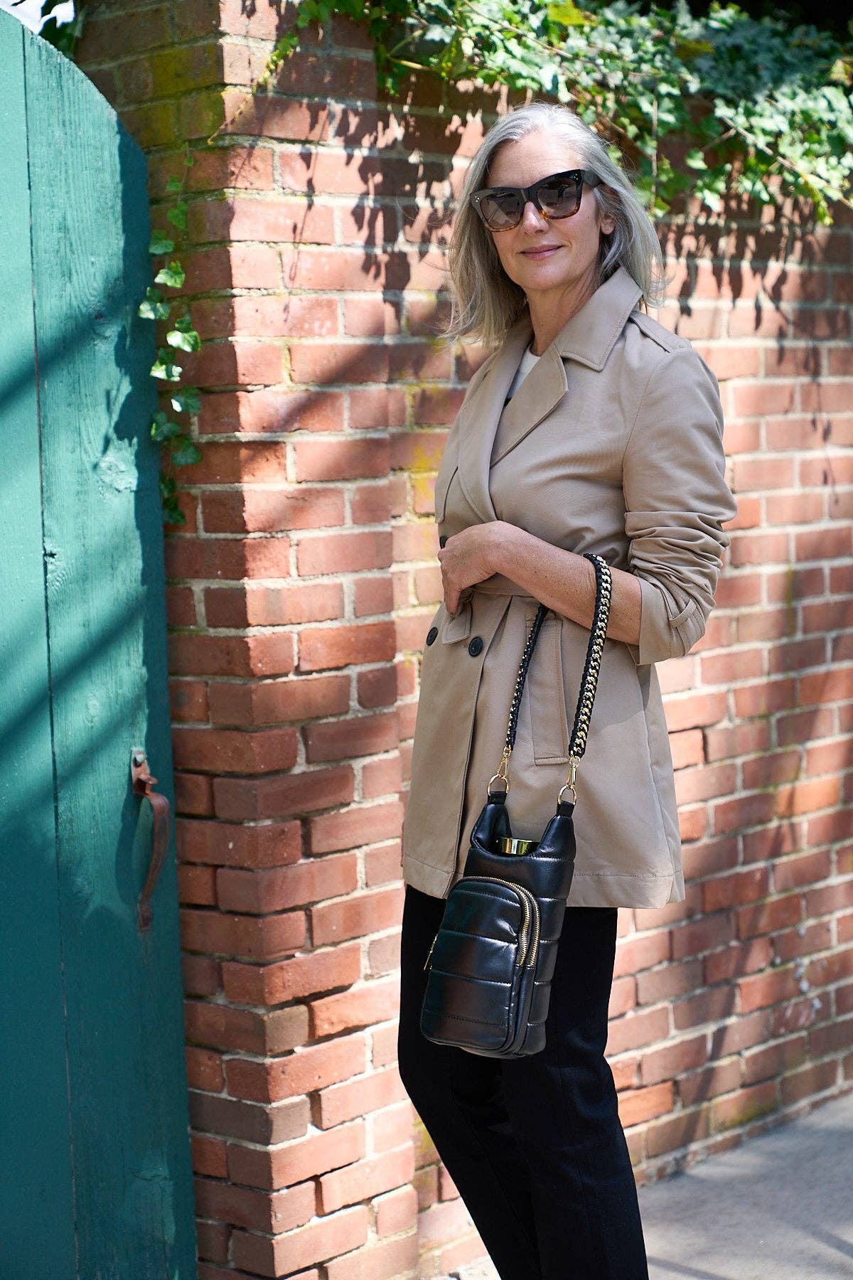 A woman with long gray hair, sunglasses, and a beige trench coat stands against a brick wall and green door. She holds the Vegan Leather HydroBag® with Strap Bundle, featuring a chic chain strap, and wears black pants. Sunlight casts shadows on the wall as greenery peeks from above.