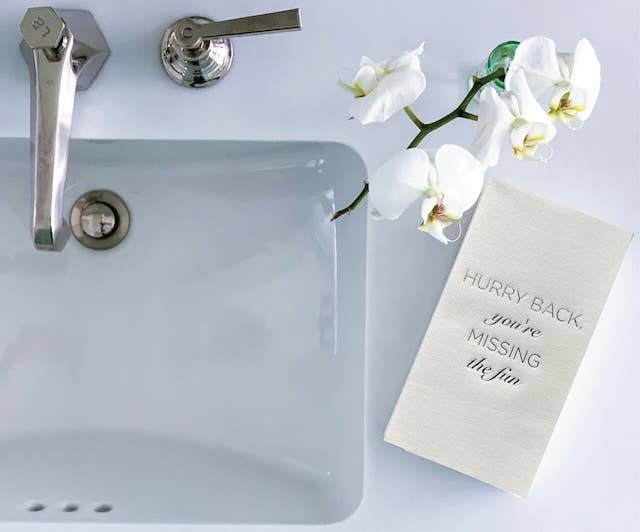 To the left is a bathroom sink with a sleek metal faucet; to the right, a white orchid with open blooms rests beside Cloth-Like Guest Towels - Year Round Themes. The towels display "HURRY BACK, you're MISSING the fun" in elegant, dark lettering.