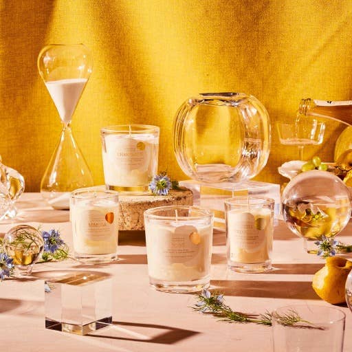 A light table is adorned with 6 oz Champagne-scented candles in glass jars with colored labels, an hourglass, a clear vase, and decorative orbs. Small blue flowers and green foliage are interspersed, all set against a mustard yellow fabric backdrop, creating a cozy ambiance.