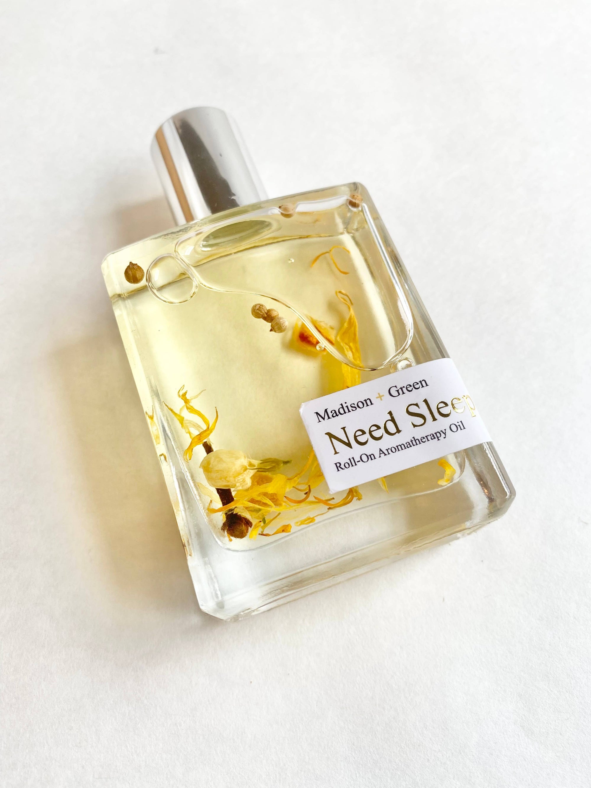 A transparent glass bottle of Madison + Green's "Need Sleep" Aromatherapy Body Oil sits on a white surface. The oil, enriched with organic essential oils and stress-relieving body properties, showcases suspended yellow and white dried flowers within its soothing fragrance. An elegant silver cap completes the sophisticated design.