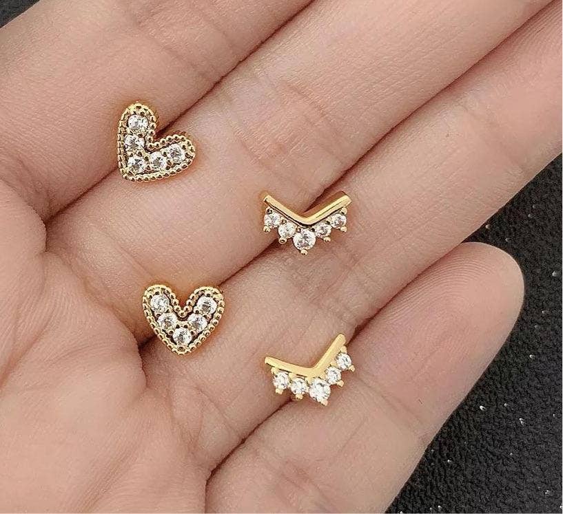 A hand showcases two pairs of 18k gold plated earrings: Nia Pave Heart Studs with embedded white stones and V-shaped earrings adorned with white stones, against a blurred dark background.