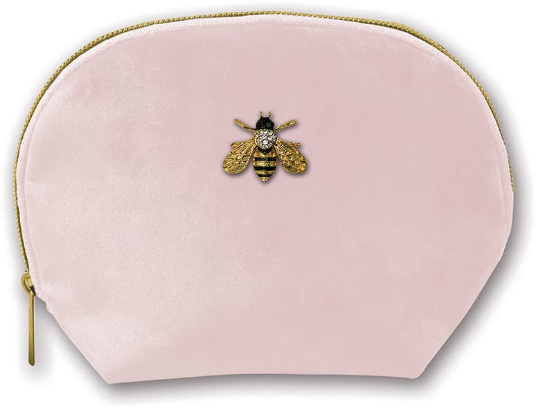 The Clamshell Bag is a light pink velvet pouch with a rounded top and gold zipper, featuring a bee brooch with jeweled body and gold wings on the white background.