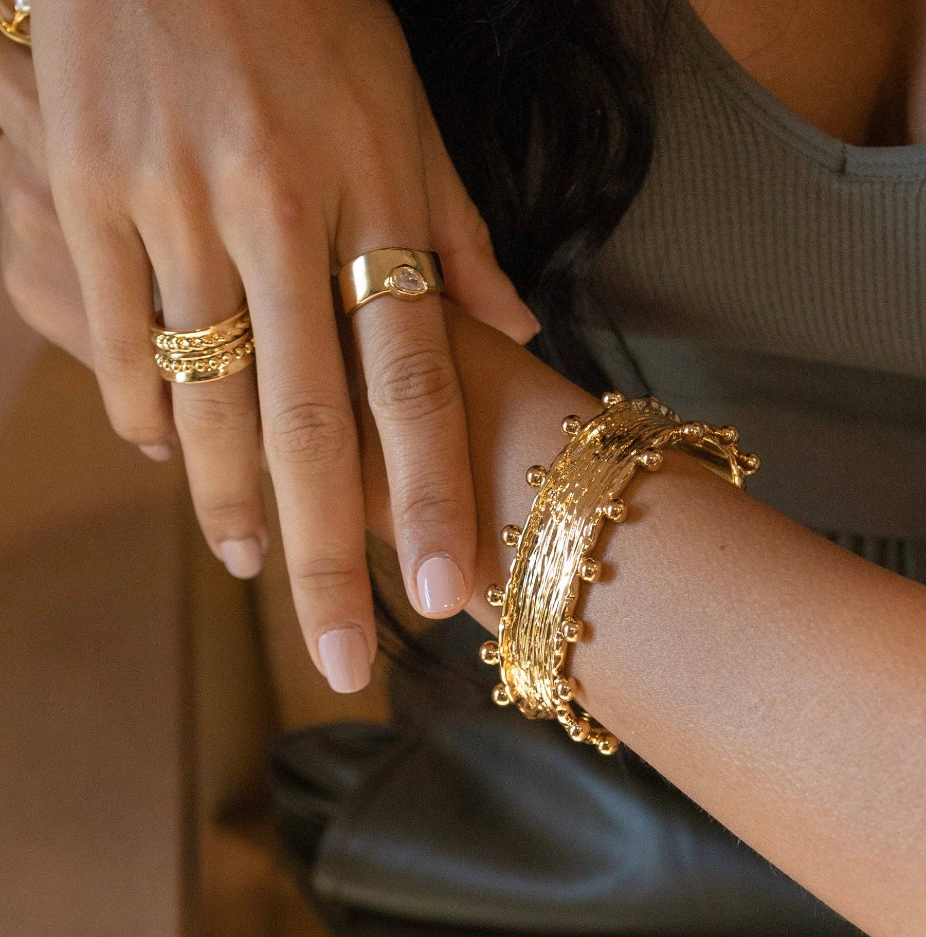 Close-up of a person wearing 18K gold plated jewelry, including a ring with a round stone on their left hand and multiple stacked rings on their right. A Raina Studded Cuff with small round accents adorns their wrist. The person wears a sleeveless, ribbed top in a neutral color.