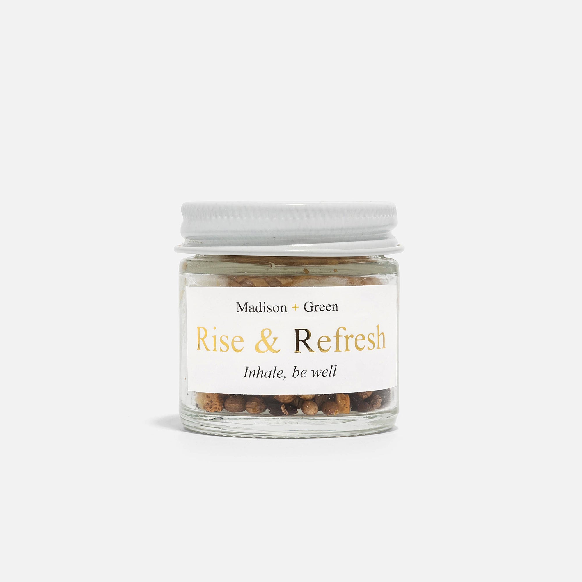 A small glass jar with a white screw-on lid, labeled "Madison + Green," features golden lettering that reads "Rise & Refresh" above the phrase "Inhale, be well." It contains a mixture of dried herbs or spices designed to blend stress-relieving scents, making it ideal for use in aromatherapy inhalers. Displayed against a plain white background, it comes as part of "The Essentials" Set of Three Aromatherapy Inhalers.