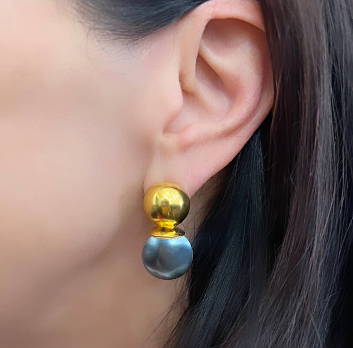 A close-up captures a woman's ear accessorized with the exquisite Gold Lady Earrings. The gold domed top sparkles brilliantly, complementing the bottom section's flat, round metallic disc adorned with a reflective silver finish. Her dark, straight hair partially veils her ear, creating an enigmatic allure.