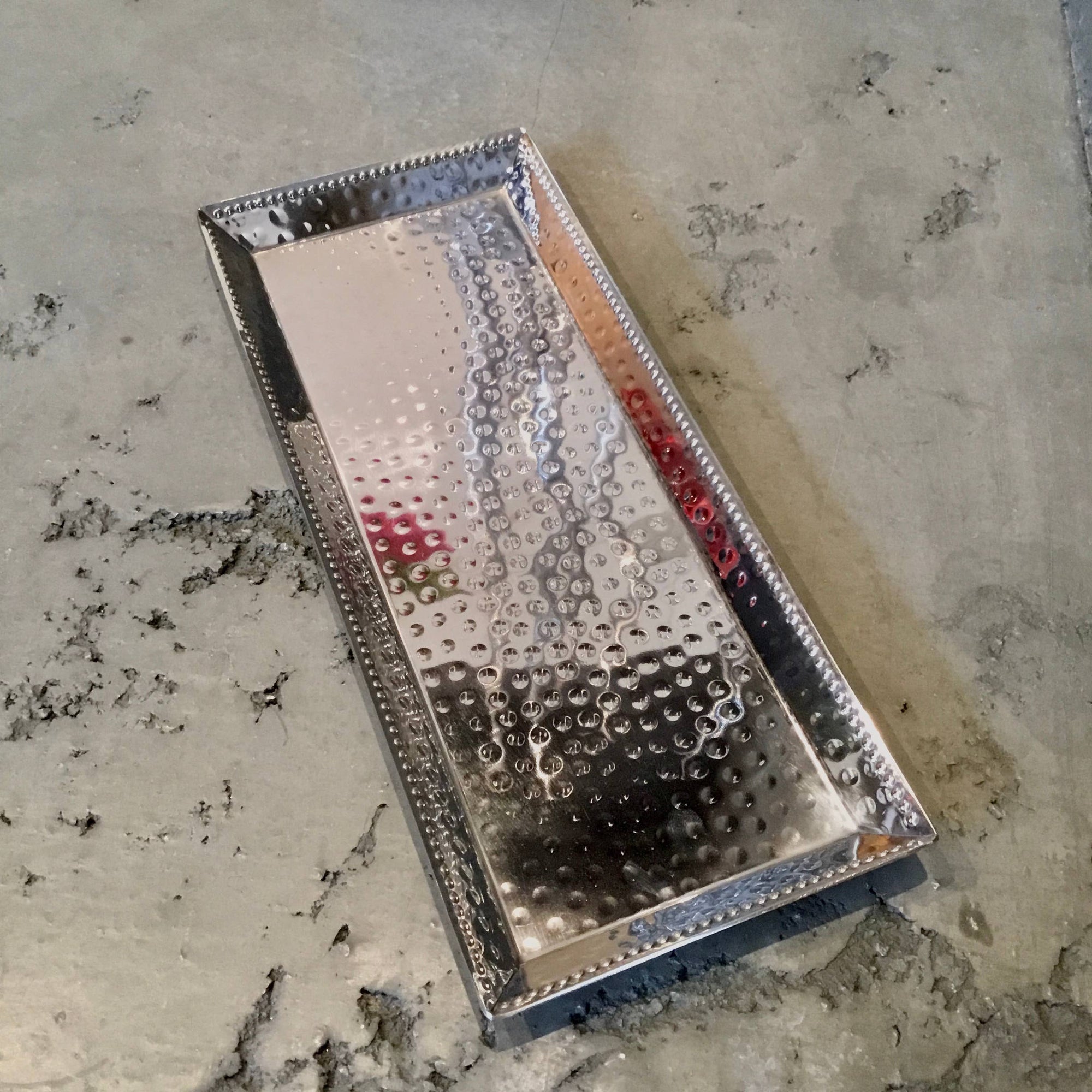 The Hammered Stainless Steel Rectangle Tray with Beaded Edges sits on a speckled gray stone surface, reflecting light off its shiny finish and dot pattern. Its edges, adorned with small indentations, add a decorative touch.