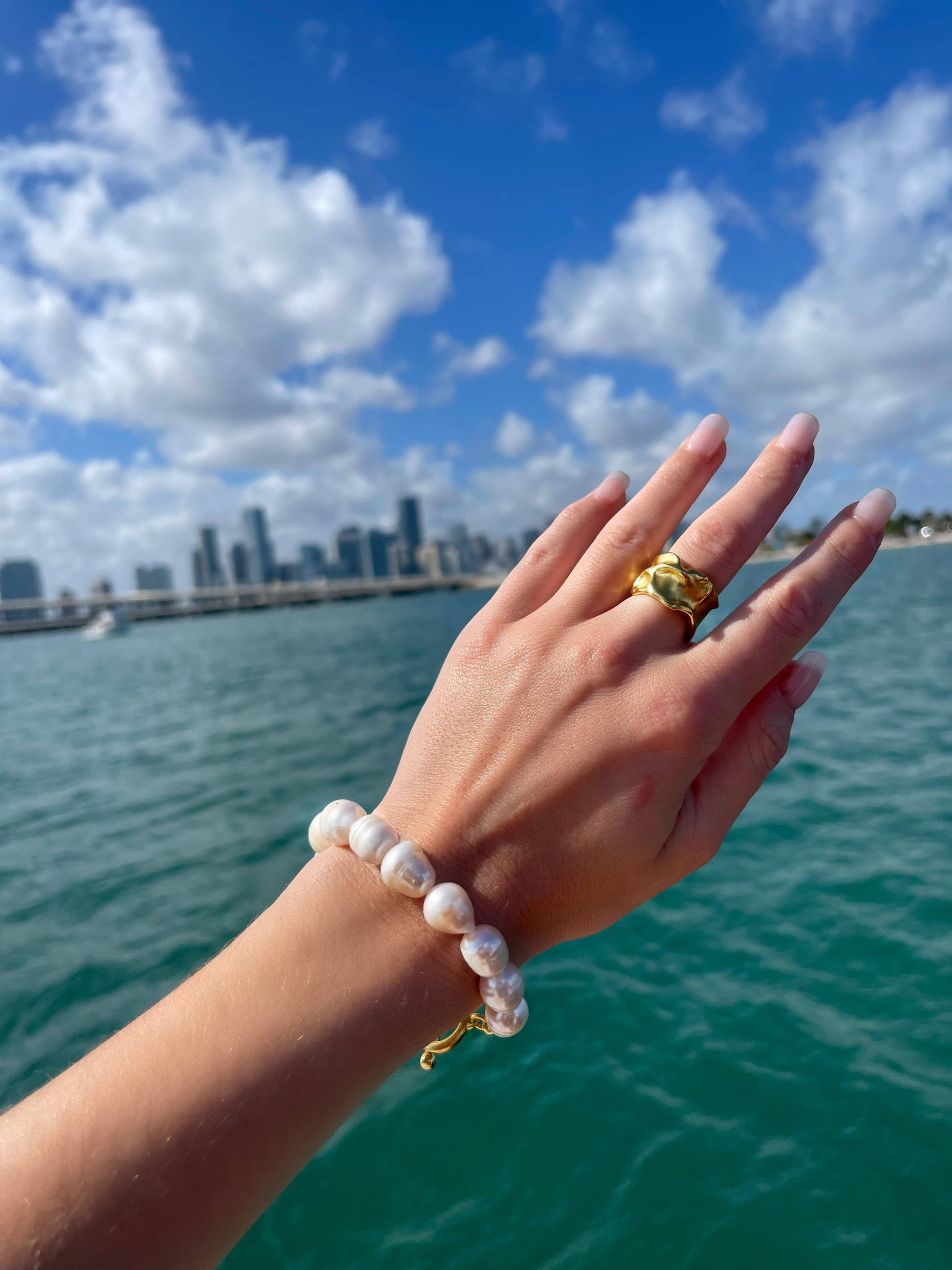 A hand adorned with a pearl bracelet and the adjustable Crumpled Foil Ring, gold-plated to perfection, is raised against a city skyline backdrop. The bright blue sky with scattered clouds mirrors in the tranquil waters below, creating a serene coastal scene.