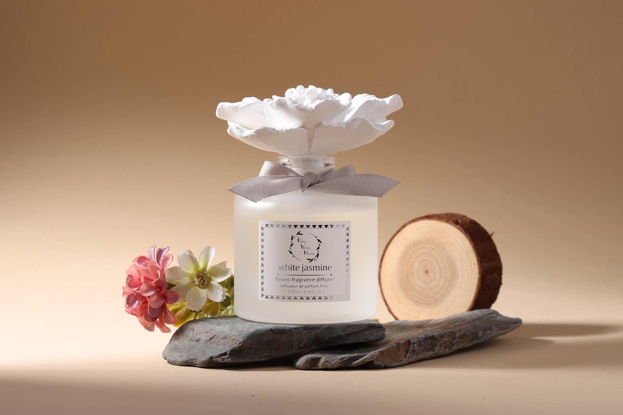 A Marigold Ceramic Flower Diffuser Gift Set - White Jasmine, featuring a frosted glass bottle adorned with a decorative white floral cap, rests on a flat stone beside an elegant box tied with a gray bow. Colorful flowers and a round wooden slice complete the scene against a smooth, warm beige background, creating a serene and sophisticated gift presentation.