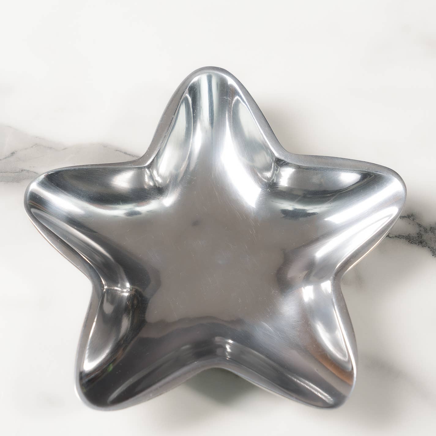 On a white marble surface with gray veining, an Aluminum Star Tray gleams with its shiny, silver finish. Its smooth, reflective surfaces and rounded edges lend it a sleek and stylish appearance. This food-safe piece requires hand washing for proper care.