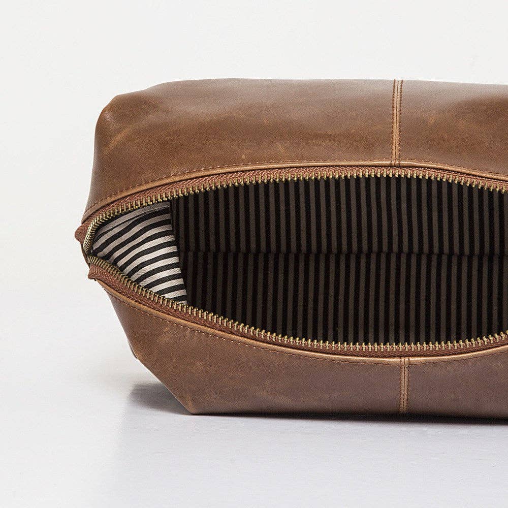 The Journeyman Dopp Kit is crafted from vegan leather in a brown hue and features a gold zipper, revealing its interior. The inside lining showcases a striking black and white vertical stripe pattern. Positioned on a neutral surface, the image emphasizes the bag's opening and design details, making it ideal for your grooming essentials.