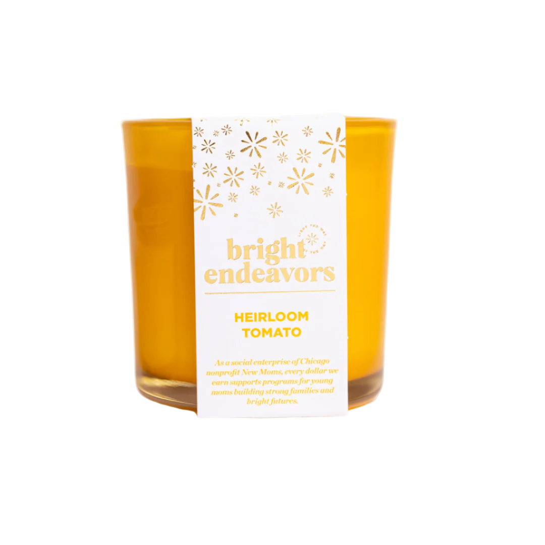 A yellow soy wax candle is nestled in a recycled glass holder featuring a label with the "bright endeavors" brand name in lowercase letters, embellished with decorative stars. The bolded text reads "HEIRLOOM TOMATO," followed by a brief description of its social enterprise mission. The fragrance is phthalate-free.