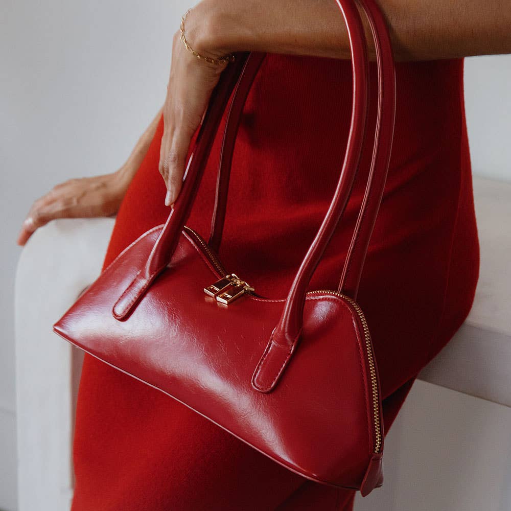 A person in a red dress holds a structured, rectangular Dakota Recycled Vegan Shoulder Bag with gold hardware. This shiny handbag rests near their hip against a soft, neutral background that contrasts beautifully with the vibrant ensemble and its OEKO-TEX certified lining.