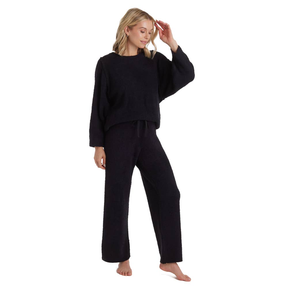 A person stands barefoot in a Marshmallow Lounge Set, one of Oprah’s favorites, exuding unmatched softness with its long sleeves and wide-leg pants. They have shoulder-length blonde hair, gently touching it with one hand while the other rests by their side against a plain white background.