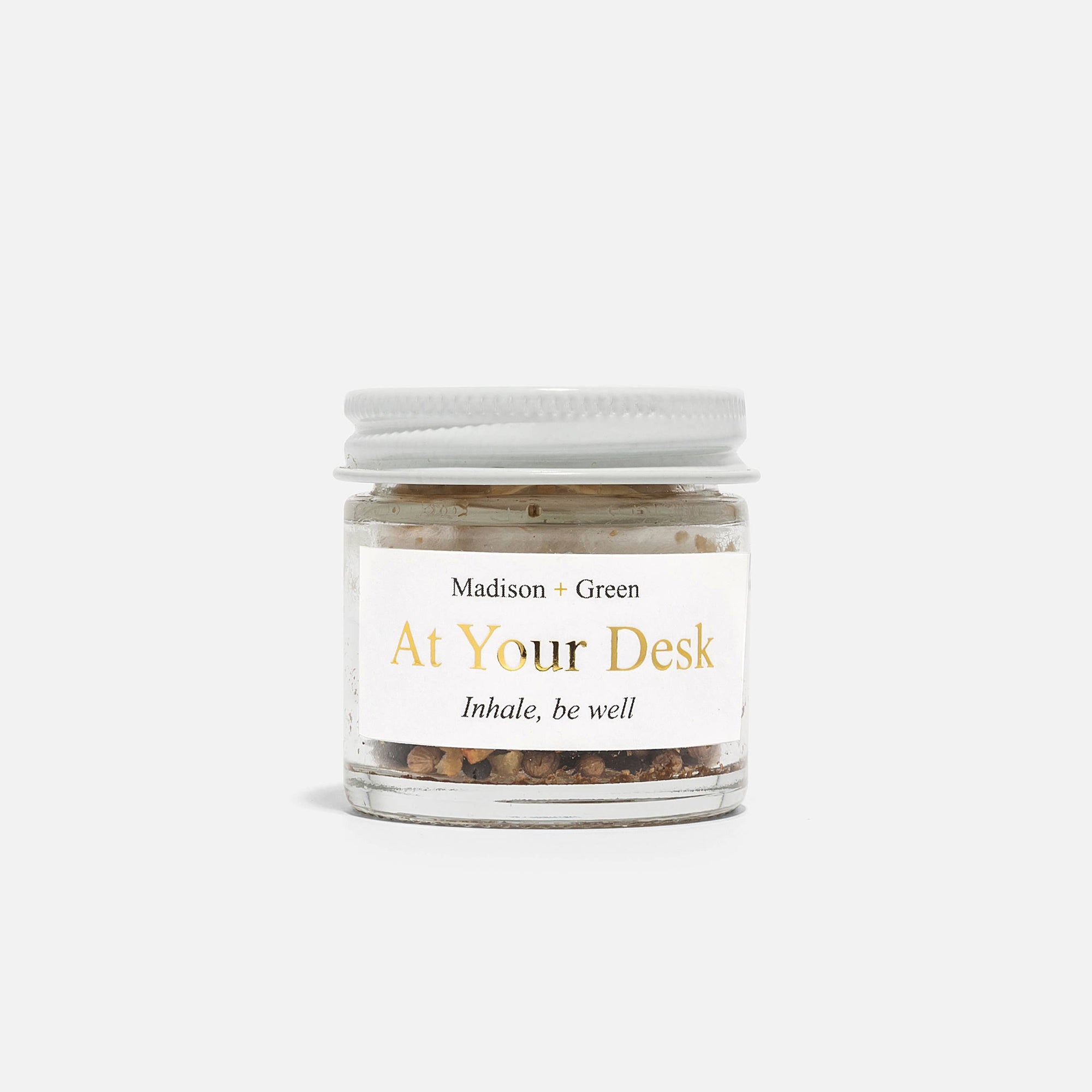 A small glass jar with a white screw-on lid contains brown granular contents, which evoke stress-relieving scents. The label prominently displays "Madison + Green" at the top and features "At Your Desk" in large print, accompanied by the phrase "Inhale, be well" underneath. This jar resembles an item from "The Essentials" Set of Three Aromatherapy Inhalers and is showcased against a simple, light gray background.