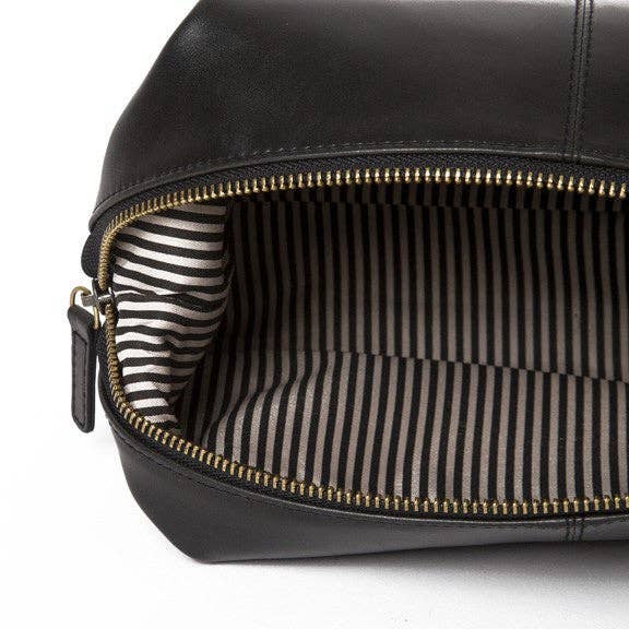 The Journeyman Dopp Kit, crafted from sleek black vegan leather, showcases a chic gold zipper that is slightly open to reveal its interior lined with striking black and white stripes. Its minimalist exterior is beautifully contrasted by the bold pattern inside, and the zipper is completed with a small leather pull tab.