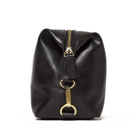 Introducing The Journeyman Dopp Kit: this black vegan leather bag boasts a vertical gold zipper down the center and features two gold rings joined by a vertical leather strap on the side. Its sleek, triangular shape and smooth finish emphasize its elegant, minimalist design.