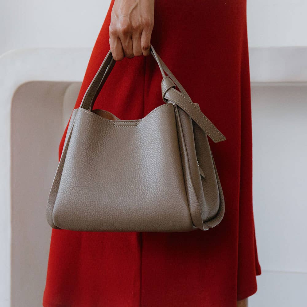 A person in a red dress holds the stylish Felicity Red Recycled Vegan Top Handle Bag, crafted from recycled vegan leather with a textured finish, wide top handle, and side flap detail. The semi-flowy dress adds elegance against an off-white background that highlights the accessories.