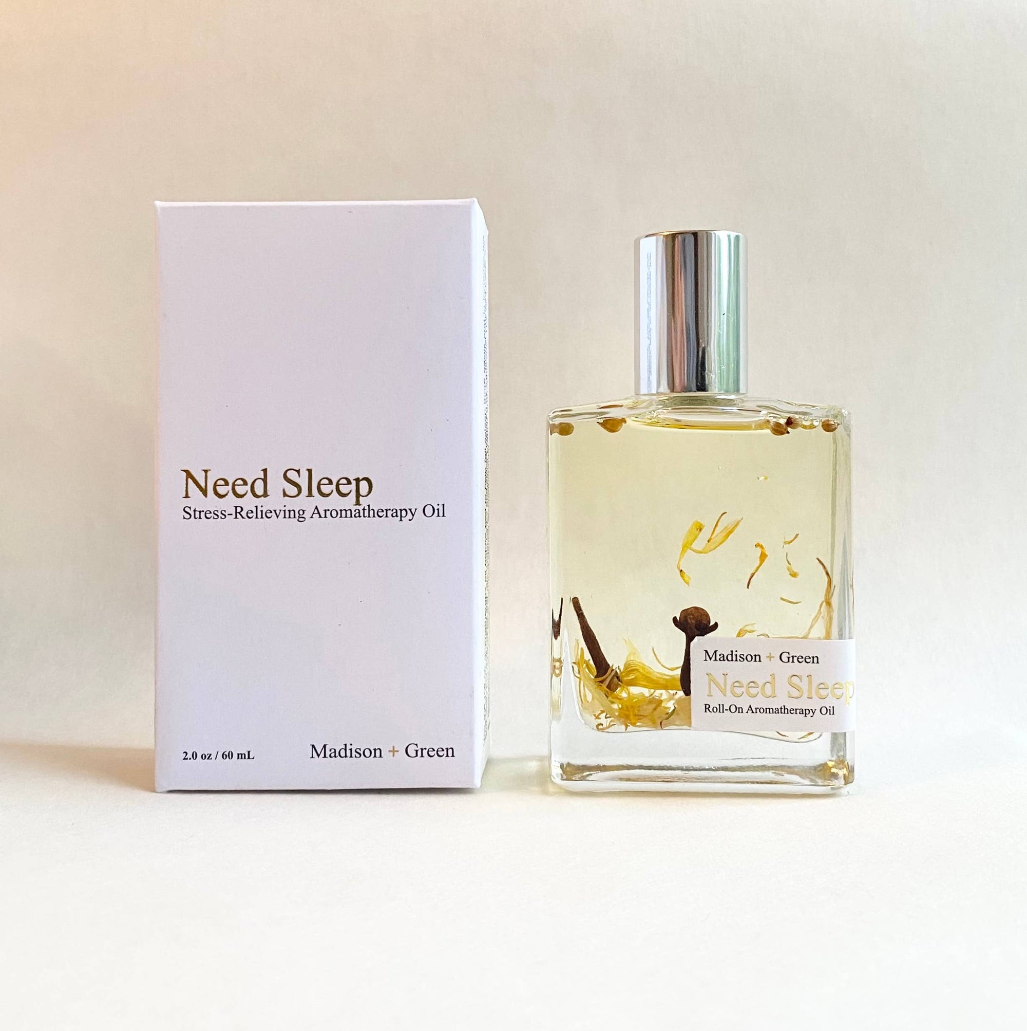 A glass bottle of "Need Sleep" aromatherapy body oil rests next to its white box. Enriched with a therapeutic aroma, the clear liquid features dried herbs. The box displays "Need Sleep, Stress-Relieving Aromatherapy Body Oil, Madison + Green, 2 fl oz / 60 ml." The bottle is capped with a metallic top.