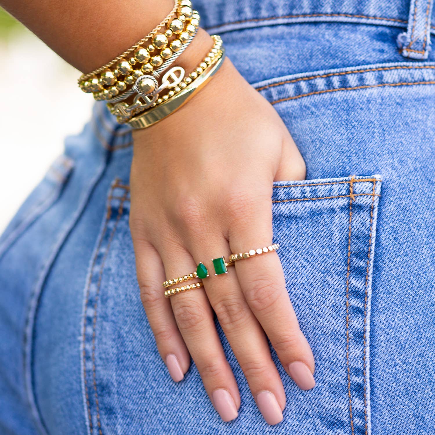 A close-up image captures a hand resting on the back pocket of light blue jeans, adorned with several gold bracelets and a modern accessory—the Gold Plated Rectangle and Pear Shaped Ring, which features small gemstones and a prominent green gem. The trendy design is accentuated by manicured nails painted with nude polish.
