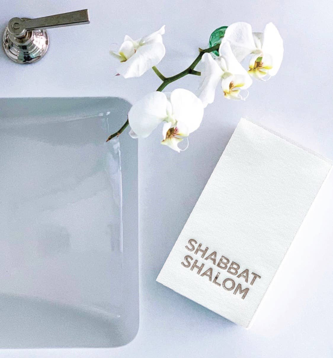 A pristine white sink with a metallic tap is on the left, with a single stem of white orchids above. To the right, a stack of foil-embossed bathroom napkins reads "SHABBAT SHALOM" in gold, adding elegance to this serene setup, ideal for welcoming guests with Cloth-Like Guest Towels - Year Round Themes.