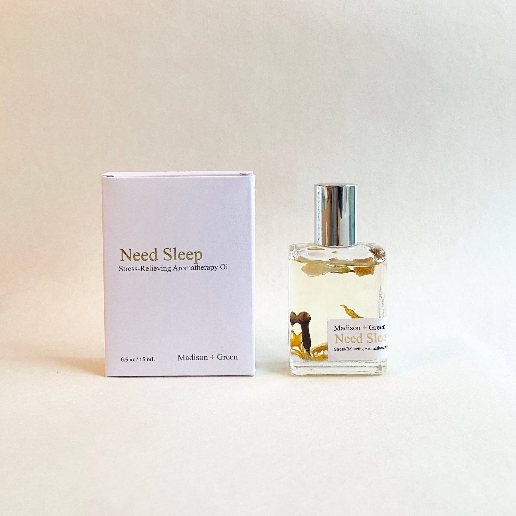 A glass bottle of the "Need Sleep" Aromatherapy Body Oil is positioned next to its white box packaging. Both the label and box prominently feature the phrases "Therapeutic Aroma" and "Madison + Green." Enriched with organic essential oils, this blend showcases delicate botanicals floating within a neutral backdrop.