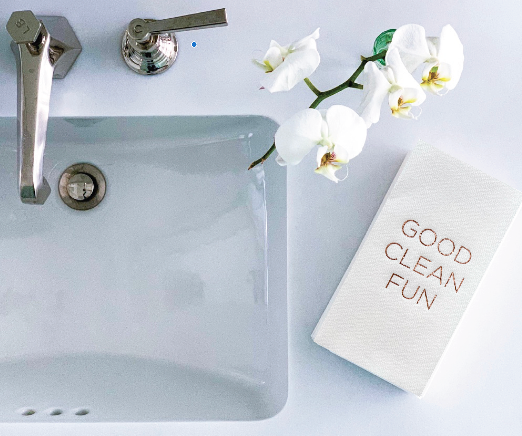 A modern bathroom countertop features a sleek silver faucet and a white ceramic sink. To the right, a white orchid branch flourishes above luxurious guest towels, neatly folded and adorned with "GOOD CLEAN FUN" in brown lettering. This scene exemplifies a clean and minimalist aesthetic using the Cloth-Like Guest Towels - Year Round Themes.