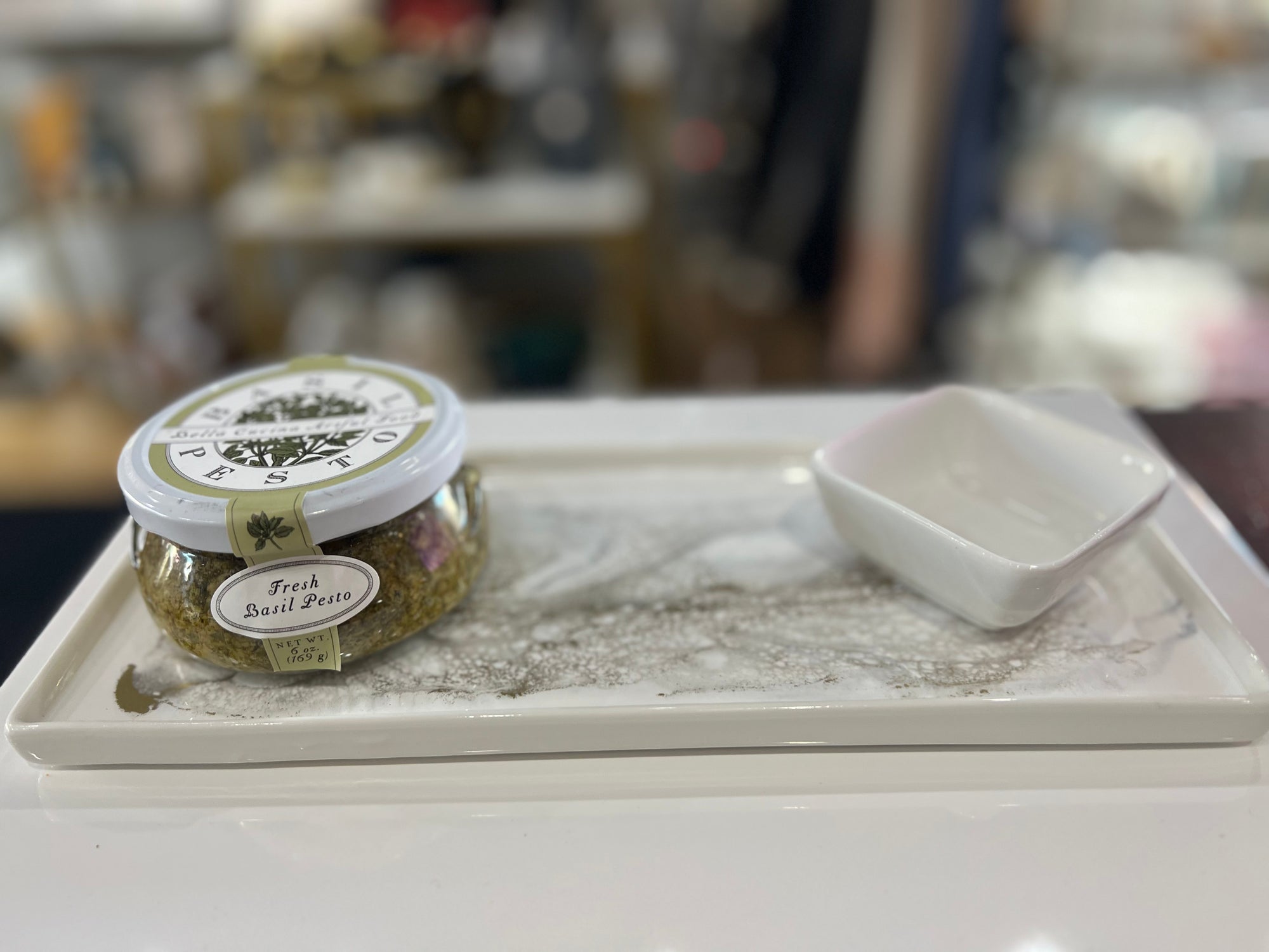 A jar of fresh basil pesto sits on a rectangular Ceramic Resin Tray, partly filled with a powdered substance. Next to the jar is a small, empty square dish. The background is blurred, showcasing an elegant interior design with various home decor items out of focus.