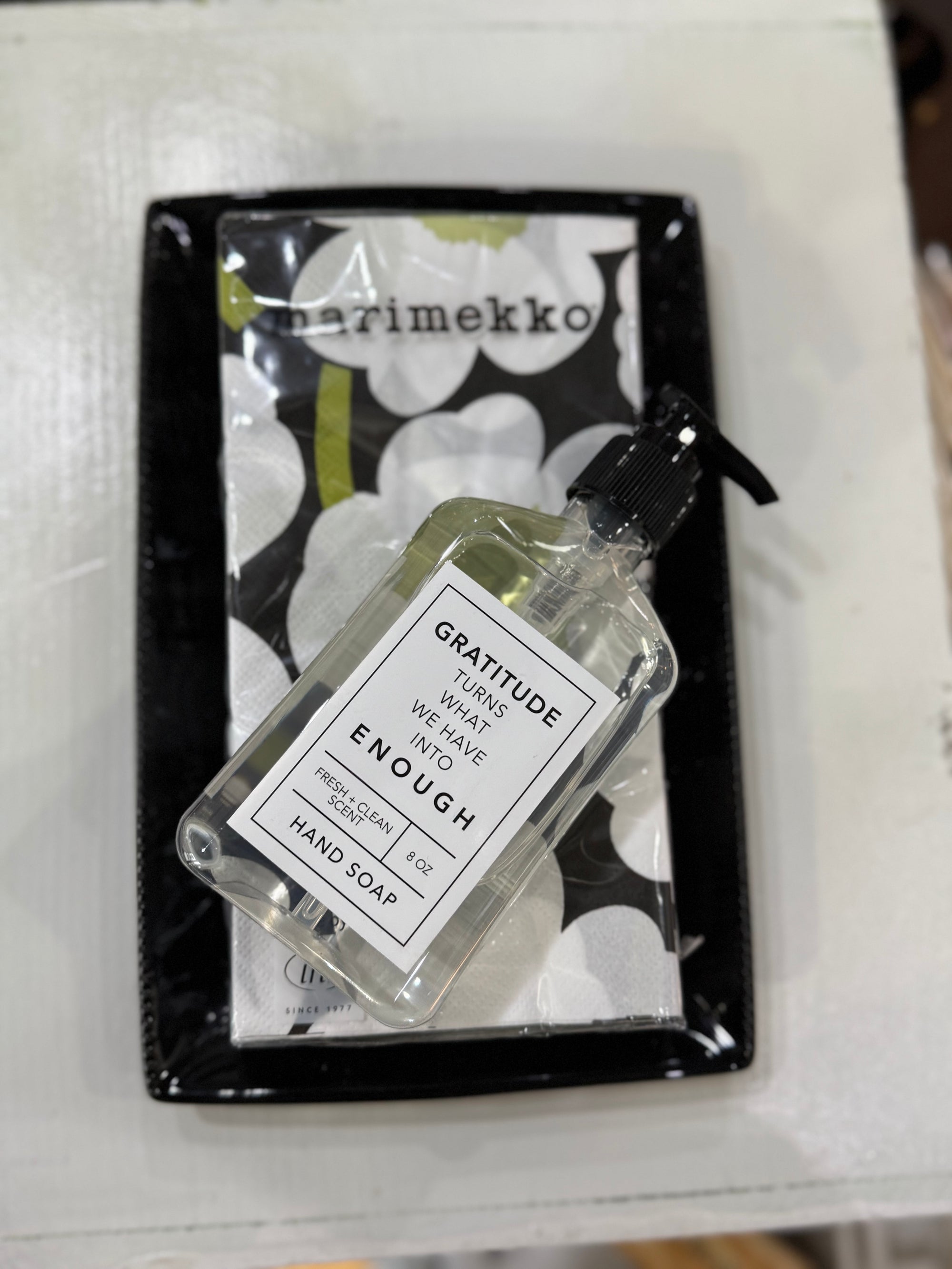 A clear bottle of Gratitude Hand Soap, marked with "GRATITUDE turns what we have into ENOUGH," is placed on a white surface, next to a package of Marimekko floral guest napkins elegantly displayed on a black tray from the Hand Towel Gift Set.