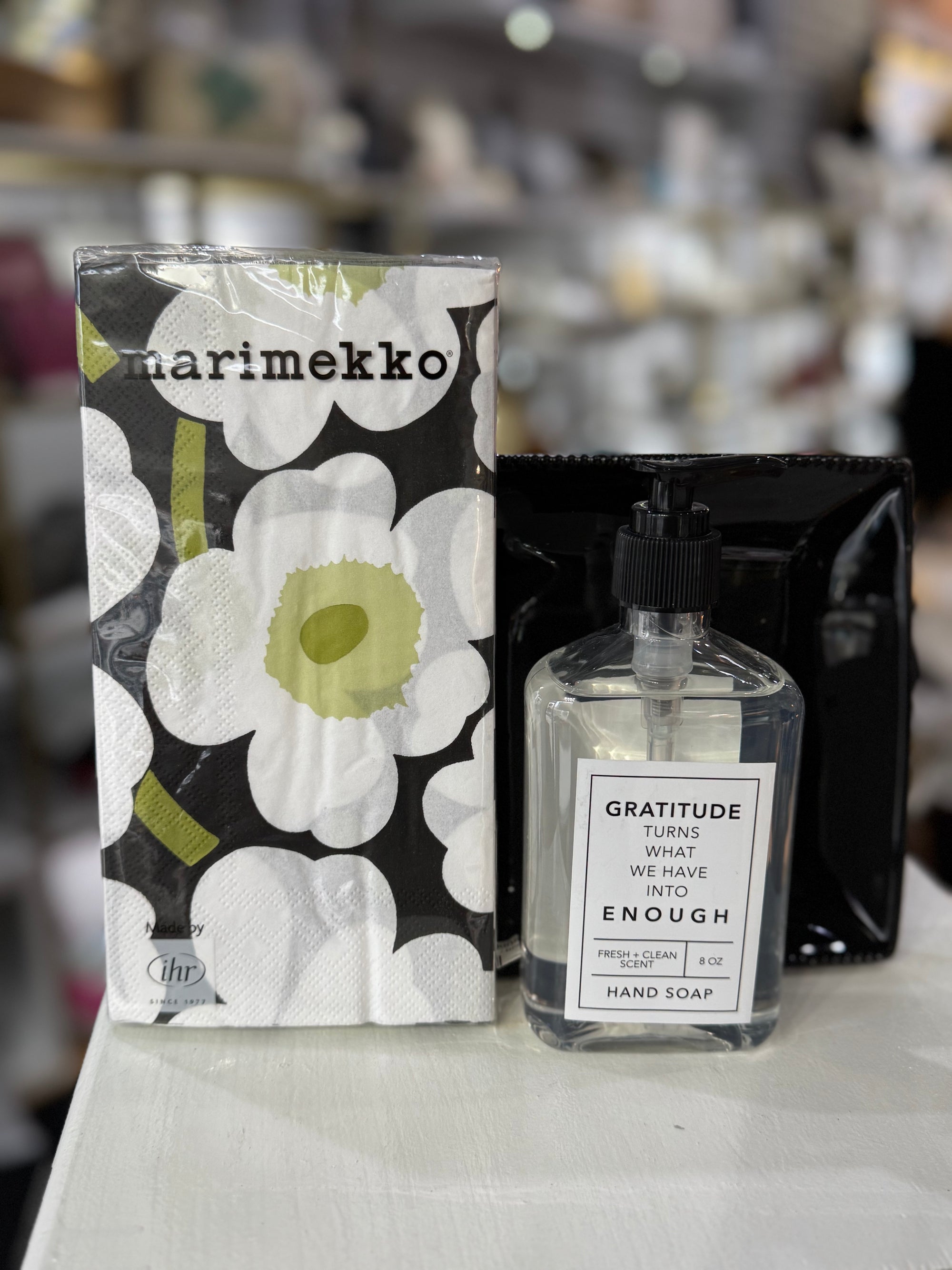 A bottle of Gratitude Hand Soap with a label that reads, "GRATITUDE turns what we have into ENOUGH," is placed next to Marimekko tissues featuring a bold floral black, white, and green design on a light-colored surface. This creates the inviting atmosphere of a Hand Towel Gift Set against a blurred store backdrop.
