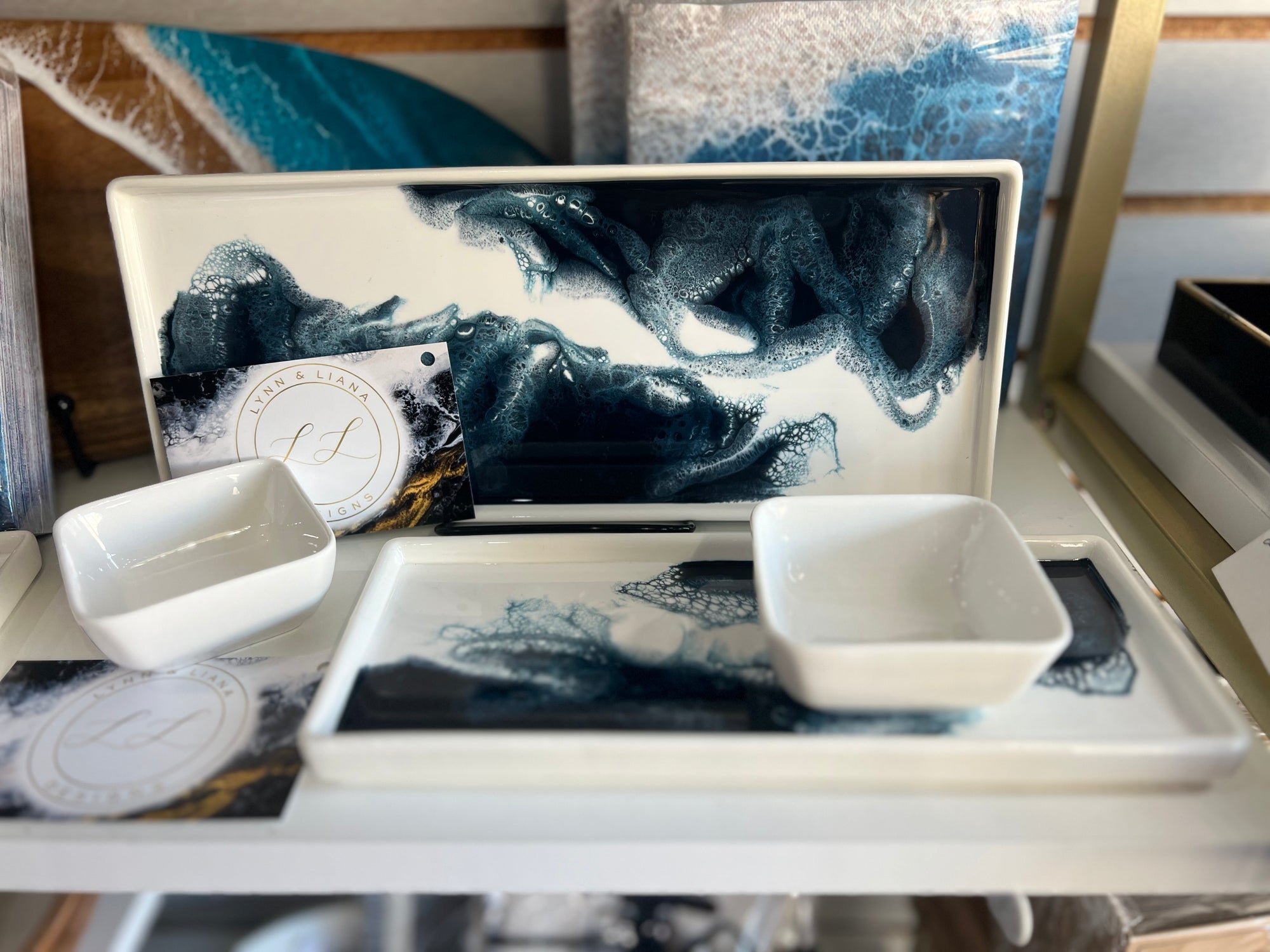 A display showcases a Ceramic Resin Tray in white rectangular form, adorned with an abstract blue and black wave design, holding two small white square dishes. Nearby, a business card with an elegant logo leans against the tray. An abstract art canvas is partially visible in the background, adding to the stylish home decor.