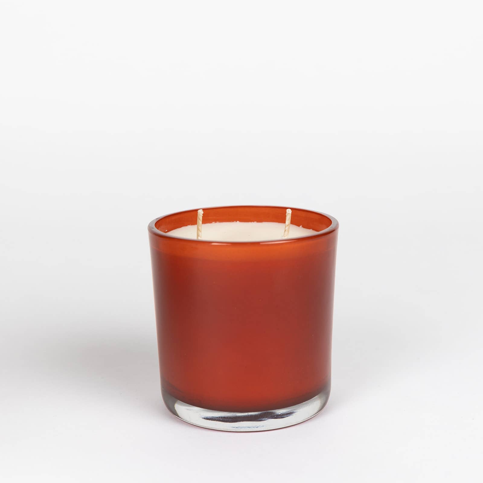 A brown glass holder contains the Pomegranate & Champagne Soy Candle, featuring a white soy wax candle with two unlit wicks, set against a plain white background. The cylindrical and translucent glass allows light to softly pass through, revealing hints of pomegranate with a phthalate-free fragrance for a pure aroma experience.
