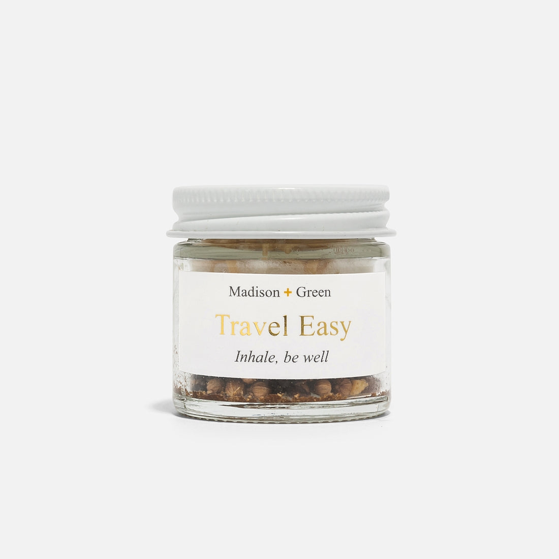 A compact glass jar with a white lid, branded as "Travel Easy" by Madison + Green, features the inscription "Inhale, be well" beneath the name. This aromatherapy inhaler showcases stress-relieving herbal or spice scents within its transparent glass, set against a plain white background.