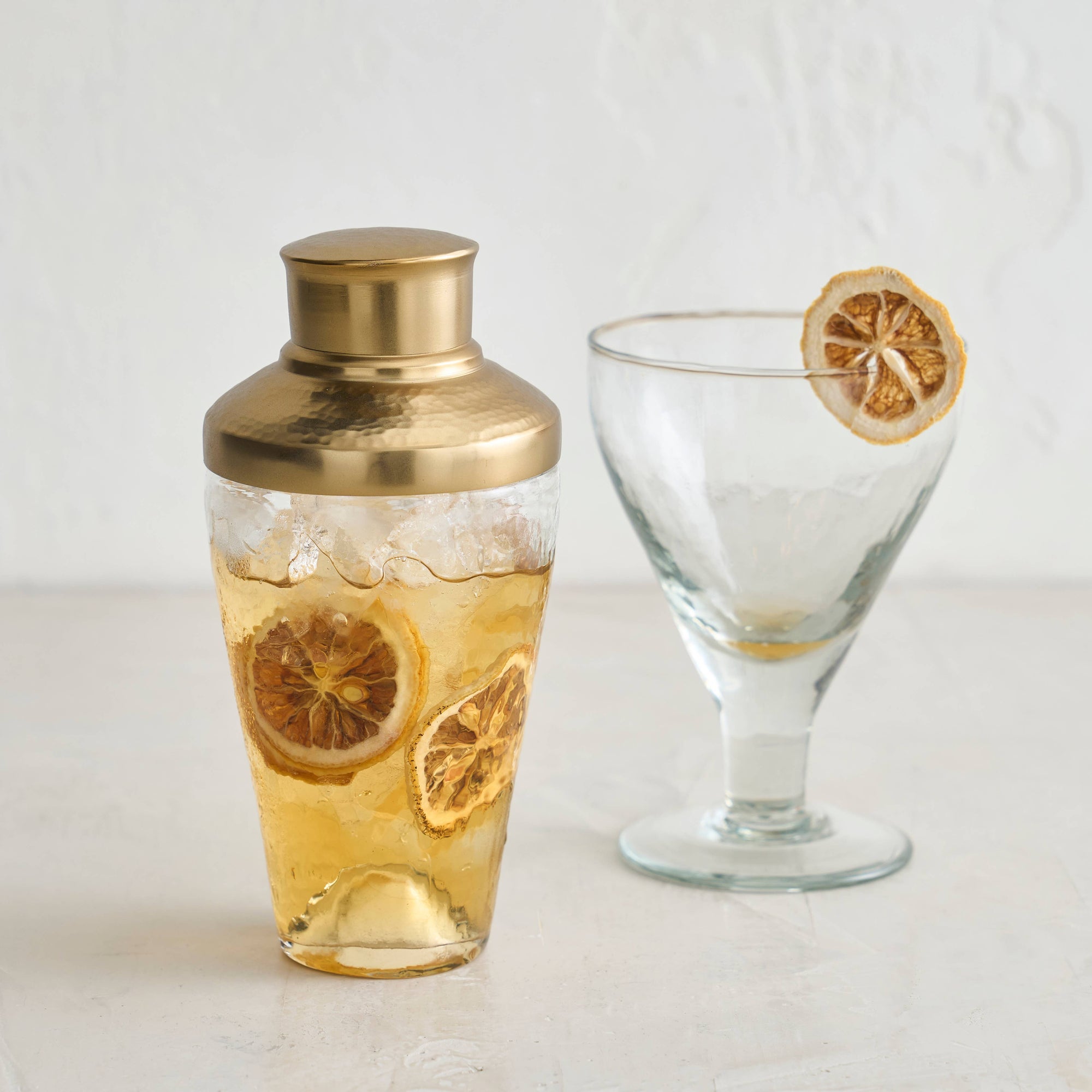 The Pebbled Mini Shaker, featuring a brass lid, is half-filled with a light brown liquid and garnished with dried citrus slices. It sits beside an empty recycled glass, also adorned with a dried citrus slice on its rim. Both are positioned on a light-colored surface against a plain background.