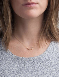 A person with shoulder-length brown hair is wearing a light gray, textured shirt and an Arch Necklace, which is a minimalist piece crafted in 14k gold-fill featuring a small curved bar. The emphasis on ethical fashion highlights the necklace and the person's neck, while keeping their facial details hidden.