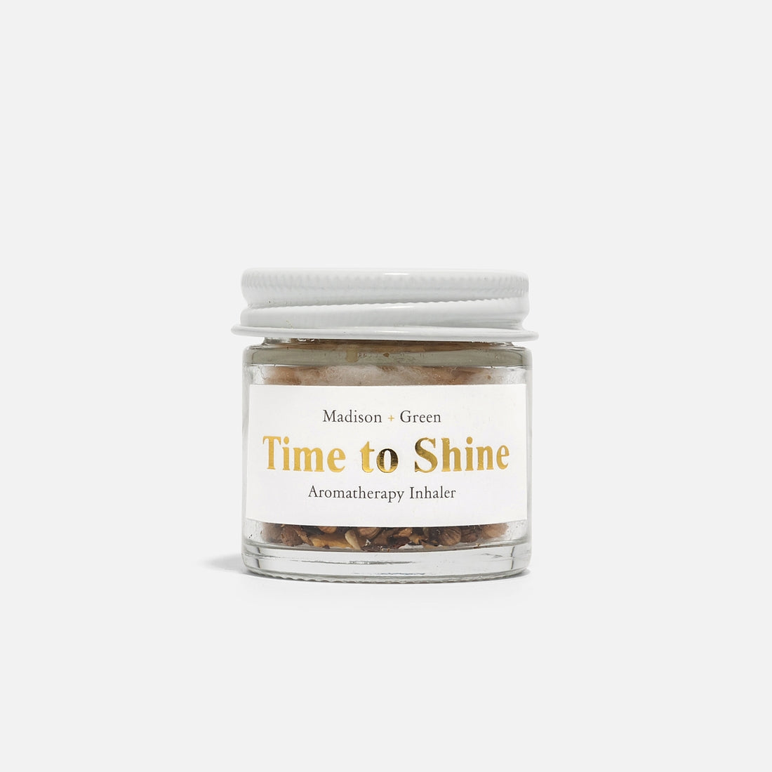 A small glass jar with a white lid showcases the label "Madison + Green" in gold text alongside "Time to Shine Aromatherapy Inhaler." Comprising a blend of organic essential oils and dried botanicals, this stress-relief fragrance is visible through the transparent glass. The background is pristine white.