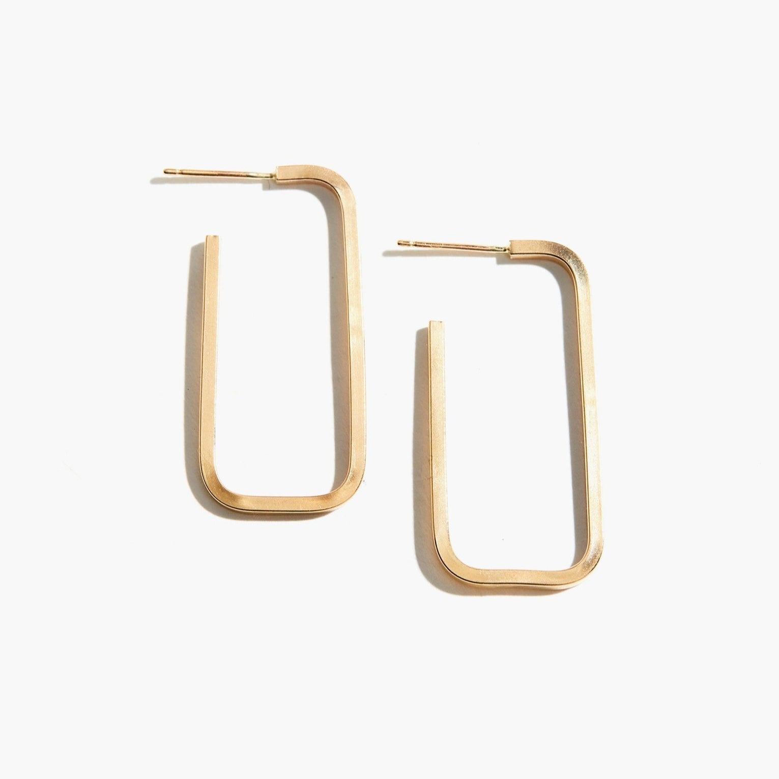 The Bali Hoops are presented against a plain white background. With their sleek and minimalistic design, each earring features an open-ended, elongated U-shape and a brushed finish, expertly crafted from 14k gold-fill. This handmade jewelry piece exudes a modern aesthetic with timeless appeal.