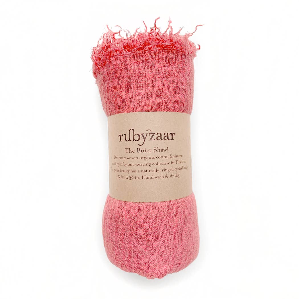 A rolled pink cotton and viscose scarf, labeled "rubyzzaar: The Boho Scarf," showcases a bohemian style. This hand-dyed piece, with frayed ends and wrapped in a beige paper band, measures 71x39 inches and recommends hand washing for care.