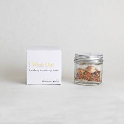 A small glass jar with a silver lid holds aromatic dried herbs, positioned beside a white box featuring the text "I Work Out" and "Stimulating aromatherapy inhaler" in grey font. This minimalist setup, designed for stress relief with essential oils, complements its light-colored background while showcasing one of the 13 varieties of Aromatherapy Inhalers.