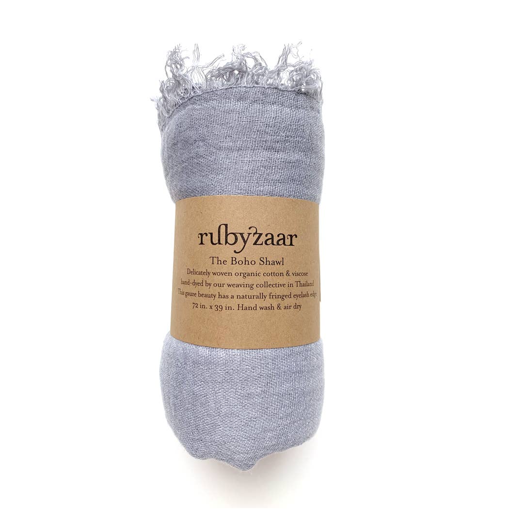 The Boho Scarf is a rolled grey shawl with fringed ends, wrapped in a brown paper band labeled "rubyzaar." Embracing a bohemian style, it is crafted from organic cotton and viscose and meticulously hand dyed. Measuring 72 inches by 39 inches, this shawl should be hand washed and air dried. It is made in Thailand.