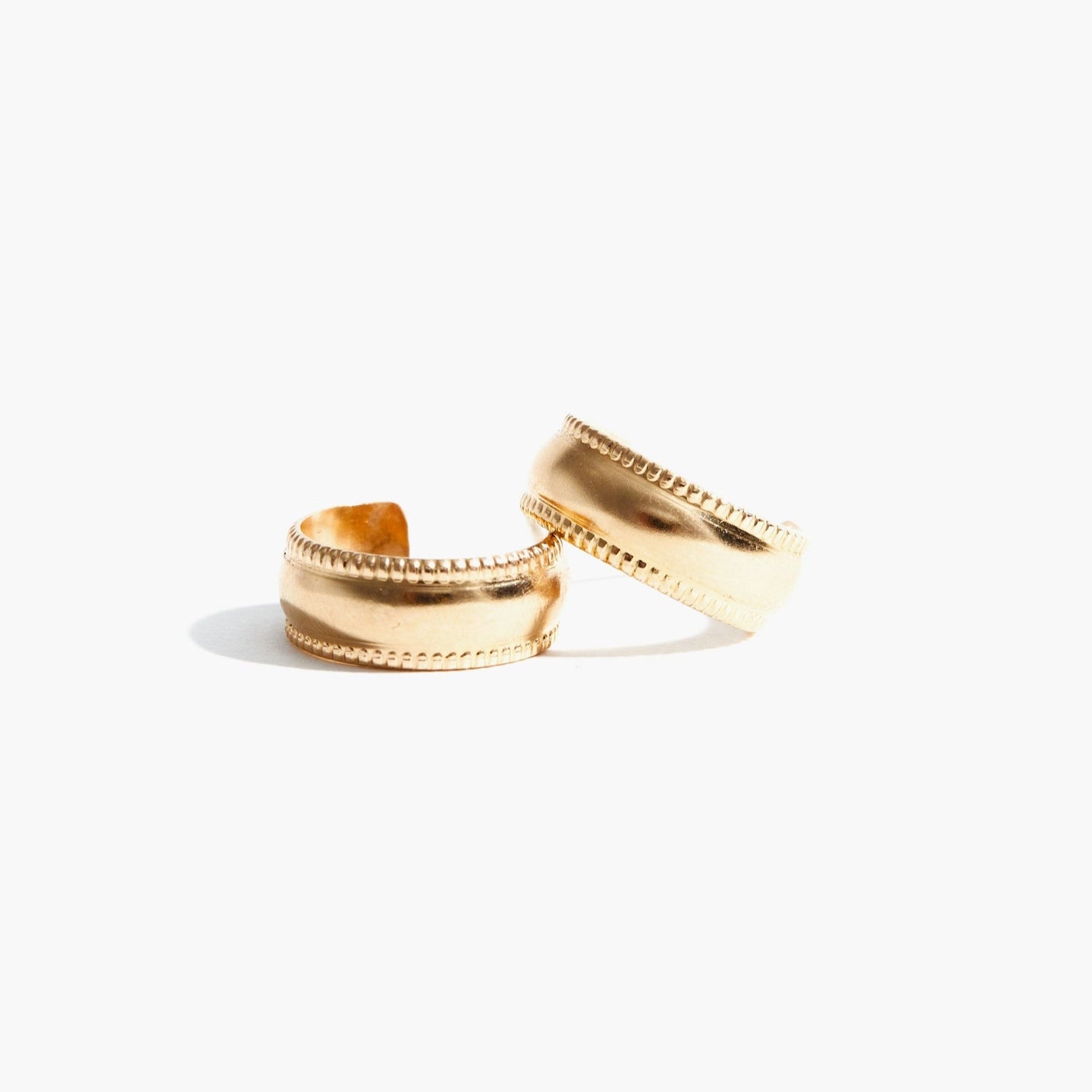 Two Chunky Column Hoops with a matte finish and delicate beaded edges are pictured on a plain white background. The positioning of the hoops suggests one is slightly leaning against the other, casting soft shadows below them—an elegant nod to an ethical fashion brand empowering women.