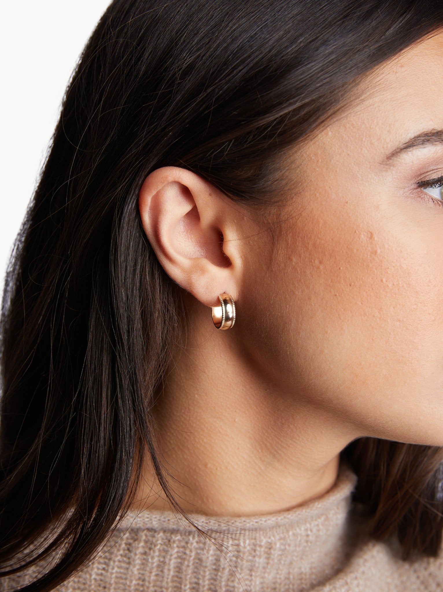A close-up image features a person with long brown hair elegantly displaying the Chunky Column Hoops, a small gold-filled hoop earring, on their left ear. They are wearing an empowering beige knit sweater against a plain white backdrop. The photo accentuates the ethical craftsmanship of the hoops, with soft shadows enhancing each intricate detail.