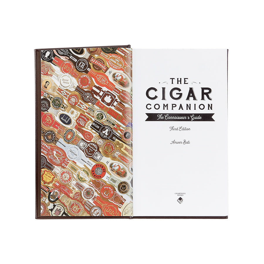 An open book titled "The Cigar Companion" (Brown Bonded Leather). On the left page, a collage of colorful cigar bands beckons cigar aficionados. The right page features the bold title text on a plain white background, celebrating the art of handmade cigars.