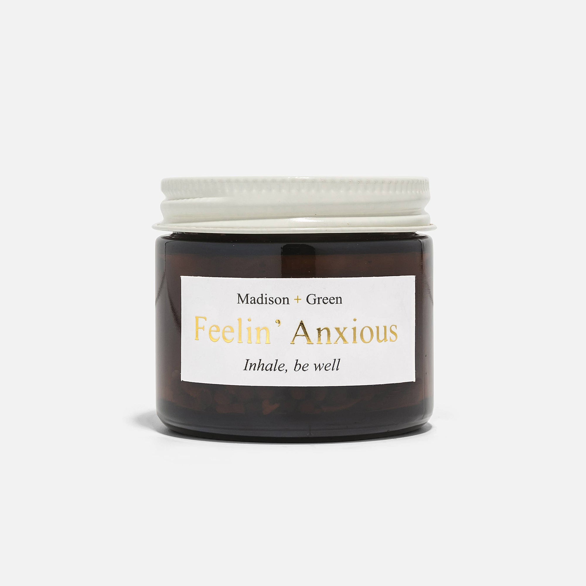 A clear glass jar featuring a white lid is labeled with "Madison + Green" and the product name "Feelin' Anxious." Designed as an aromatherapy inhaler to help alleviate stress, it carries the inspirational text "Inhale, be well," suggesting its contents are infused with essential oils. The jar is showcased against a simple white backdrop.