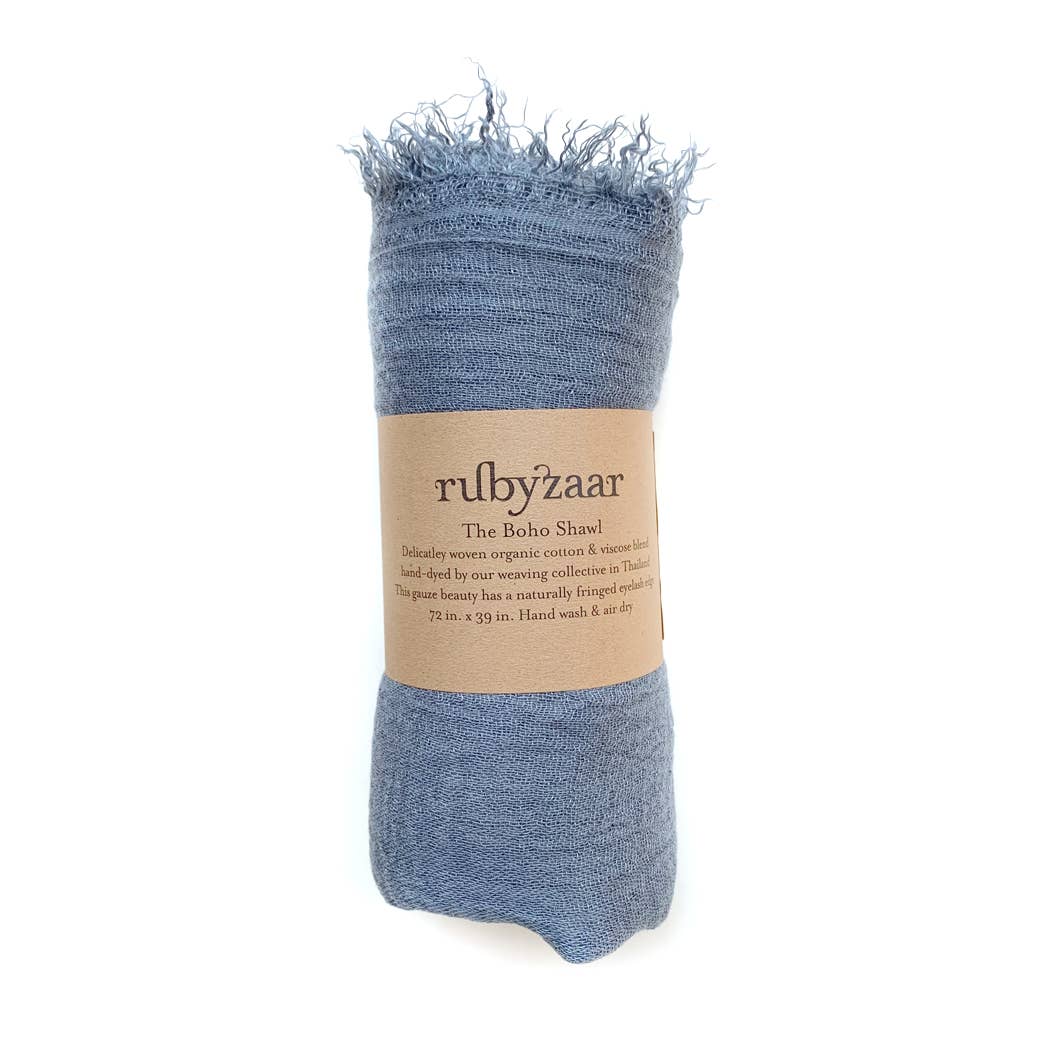 A rolled-up blue Boho Scarf with frayed edges is secured by a brown label that reads "rubyzaar, The Boho Shawl." This bohemian-style scarf is delicately woven from a blend of cotton and viscose fibers. It features hand-dyed, earth-friendly colors. Measuring 72 x 39 inches, it requires hand-washing only.