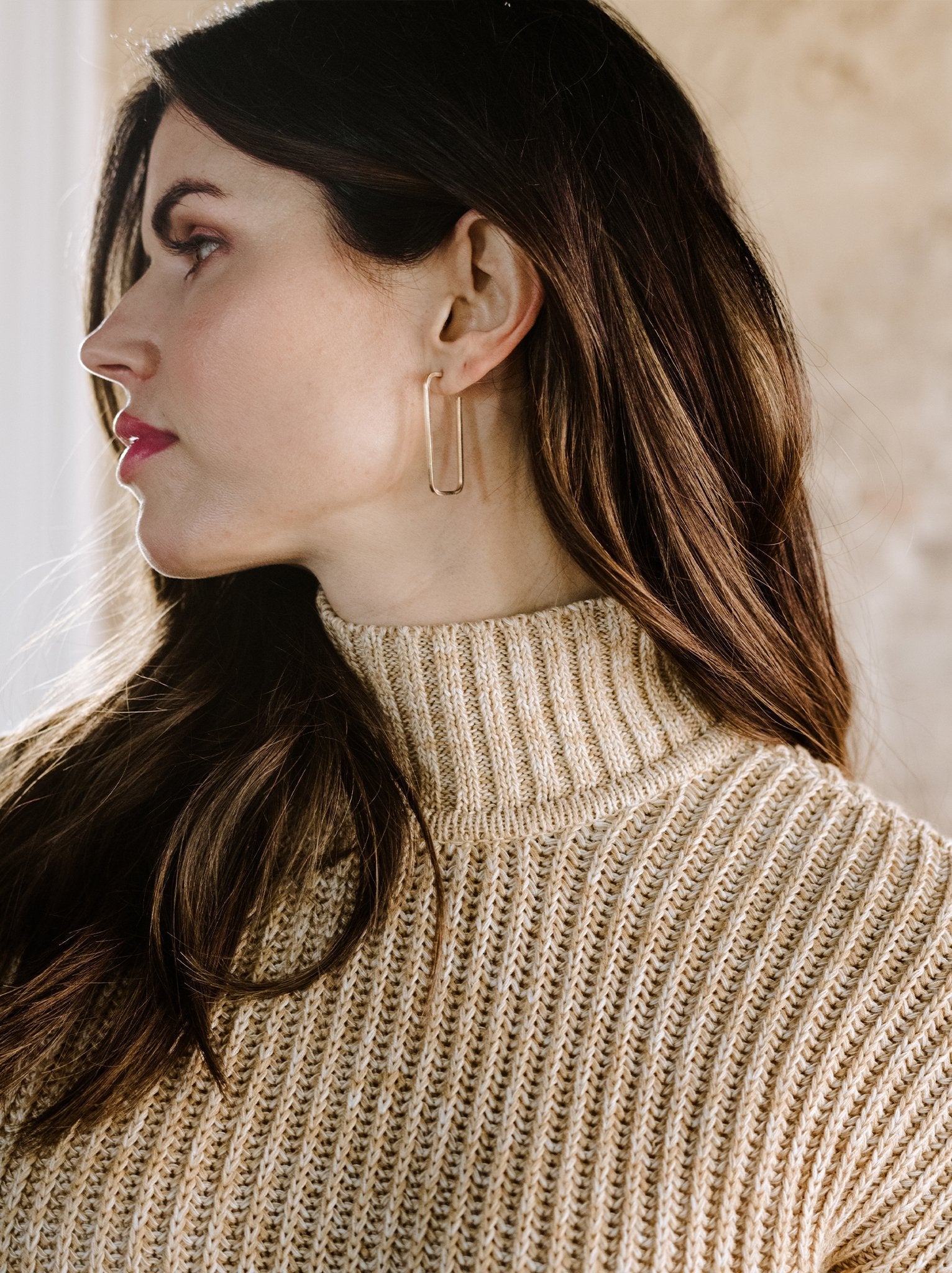 A woman with long brown hair is shown in profile, wearing a beige knit turtleneck sweater and Bali Hoops crafted from 14k gold fill. Her expression is calm as she gazes slightly upwards, set against a softly blurred background that creates a warm, neutral tone.