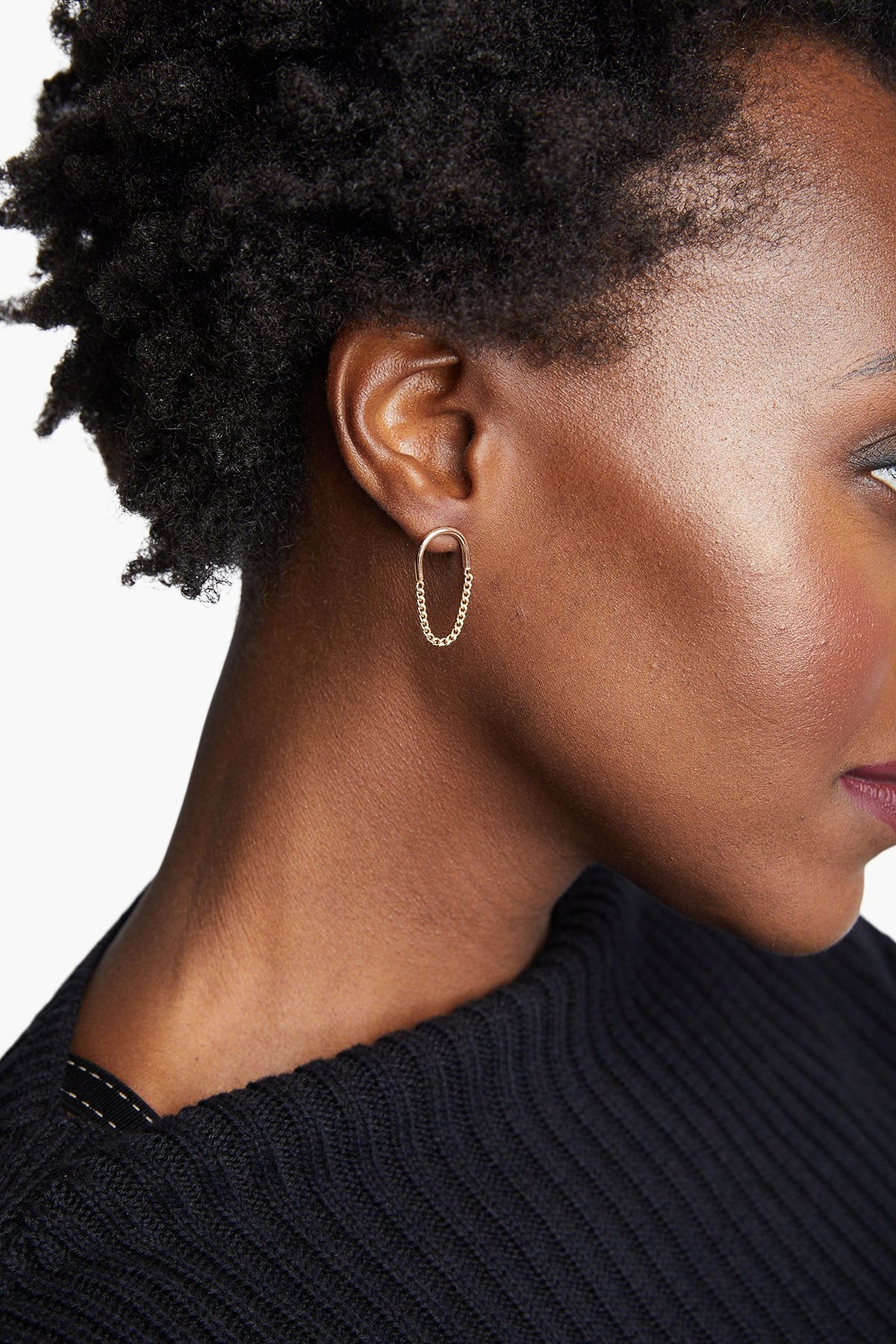 A person with short, textured hair is shown in profile wearing a black ribbed sweater. They have an oval-shaped gold-filled earring, known as the Arc Chain Earrings, embodying ethical fashion values. The white background highlights the elegant contrast with their skin tone and the striking earring.