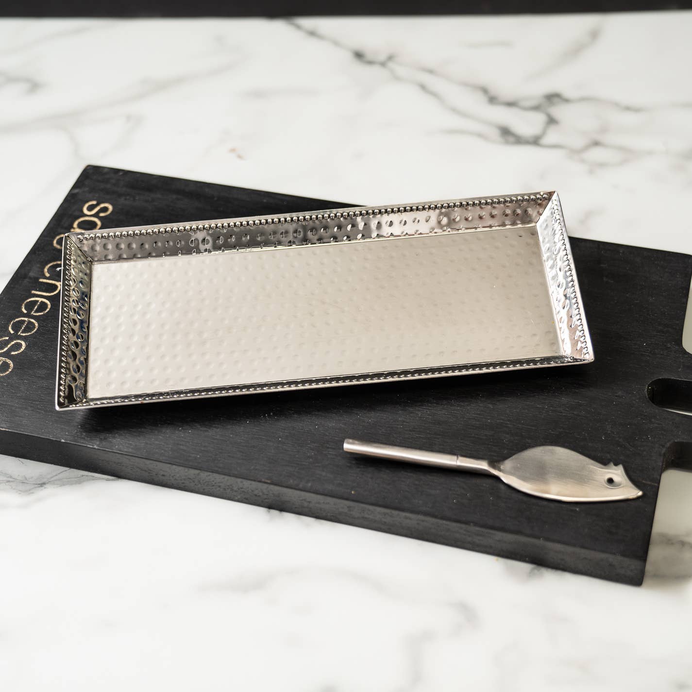 A Hammered Stainless Steel Rectangle Tray with Beaded Edges is elegantly placed on a black cheese board, which partially reveals the word "cheese." The setup rests on a white marble surface, accompanied by a small silver cheese knife featuring a leaf-shaped blade beside the tray.